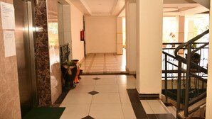 A hotel lobby with a marble reception desk, a fire extinguisher, a staircase with a metal railing, and a tiled floor. - Sherry Homes Raha Westlands (Nairobi)