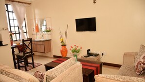 A living room with two sofas, a coffee table, a dining table with chairs, a TV mounted on the wall, and a vase with flowers on the coffee table. - Sherry Homes Raha Westlands (Nairobi)