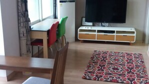 A living room with a wooden floor, a dining table with chairs, a TV on a white stand, and a patterned rug. - Tongyeong Camellia House (Tongyeong)