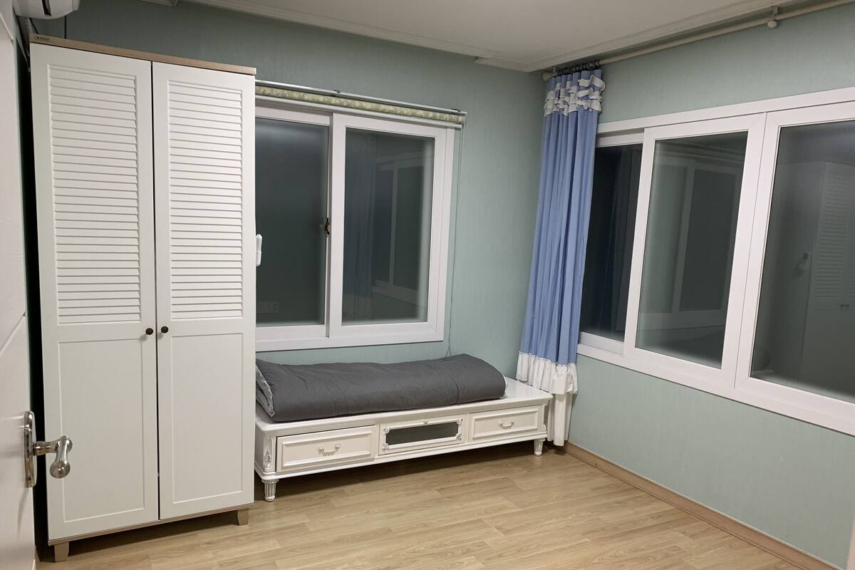 A room with a bed, a wardrobe, a window with curtains, and a wooden floor.