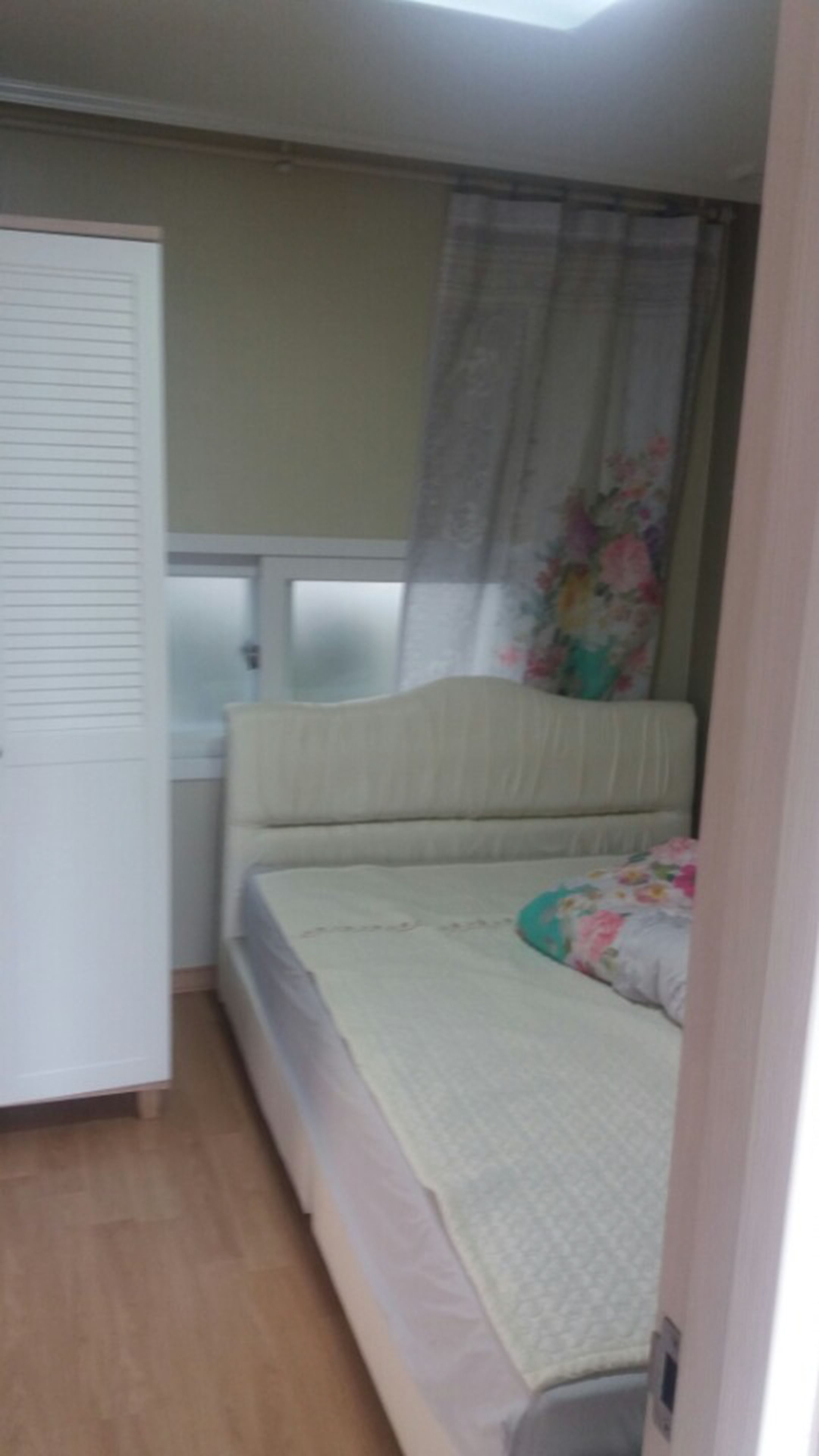 A small, single-bed room with a wooden floor, a white wardrobe, a window with a floral curtain, and a bed with a quilted cover.