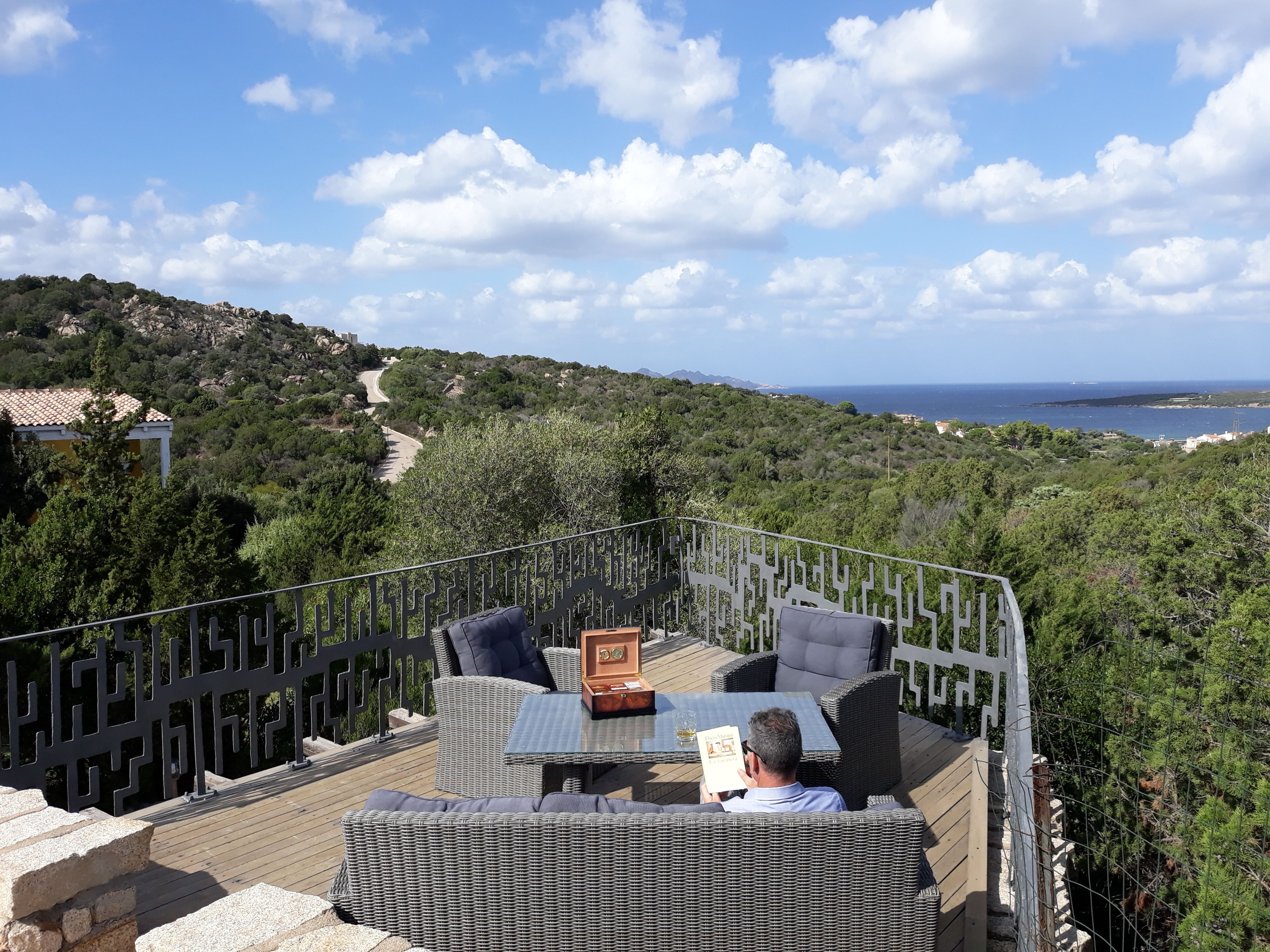 A terrace with wicker furniture, a table, and a view of the sea and hills.