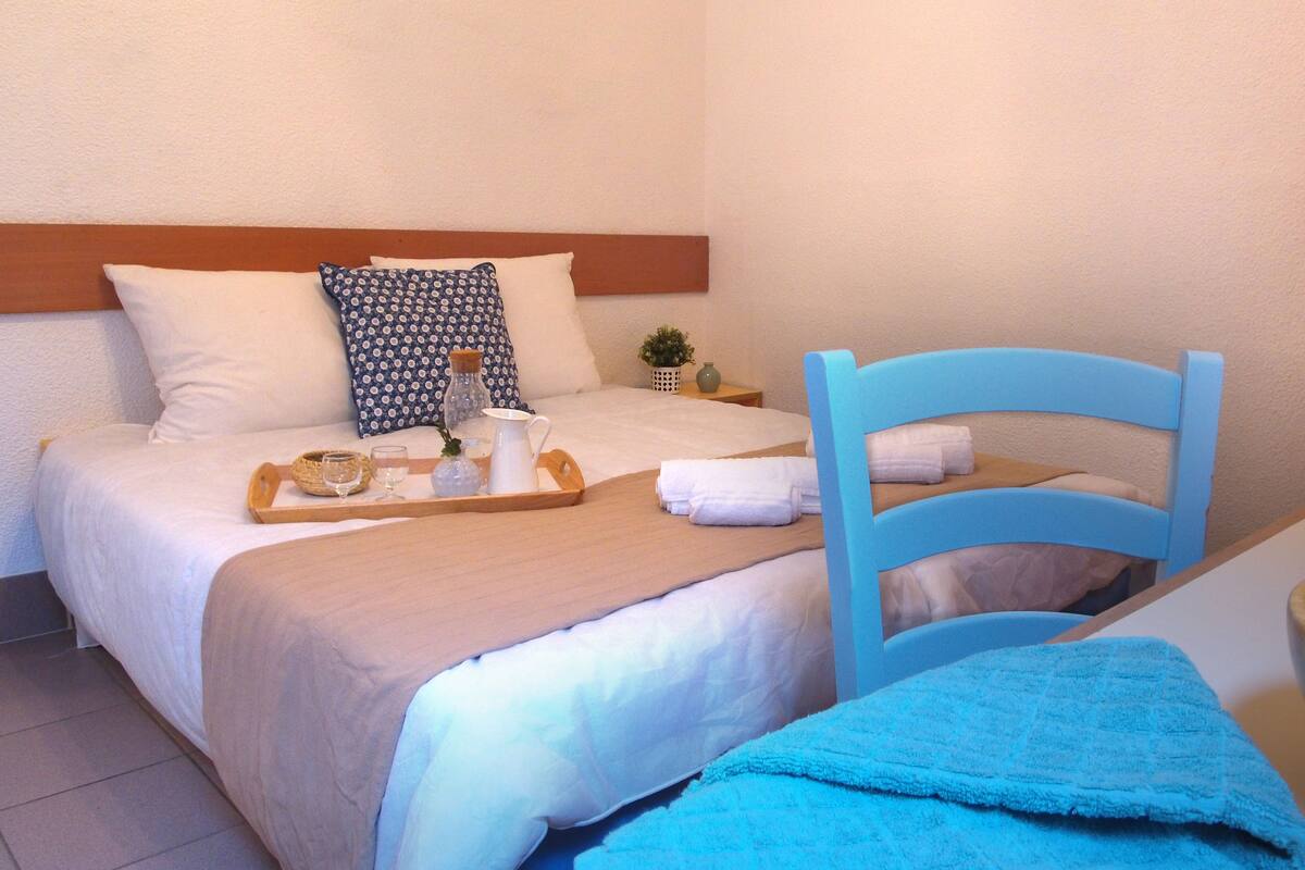 Un llit ben fet amb un safata de menjar, una cadira blava i un bol de fusta damunt d'una taula.