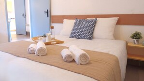 Ein ordentlich gemachtes Bett mit weißen Leinentüchern, einem Holzkopfende und einem blau gemusterten Kissen. Auf dem Bett liegen zusammengerollte Handtücher und ein Tablett mit einer Flasche und Gläsern.