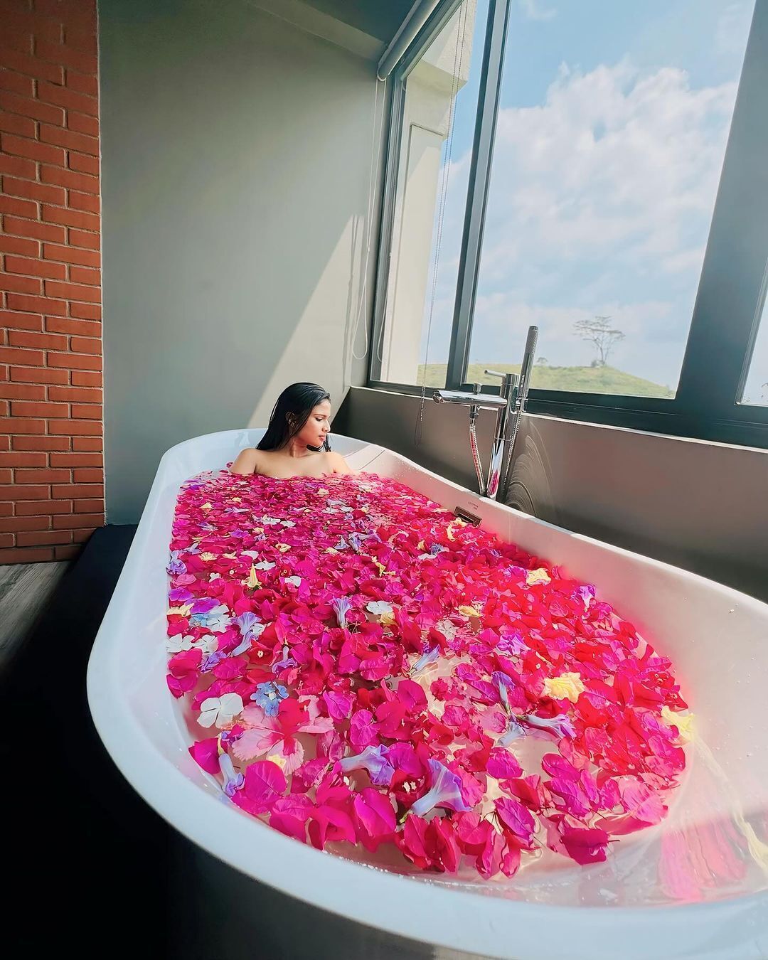 Una vasca da bagno piena di petali di rosa, una finestra e un muro in mattoni.
