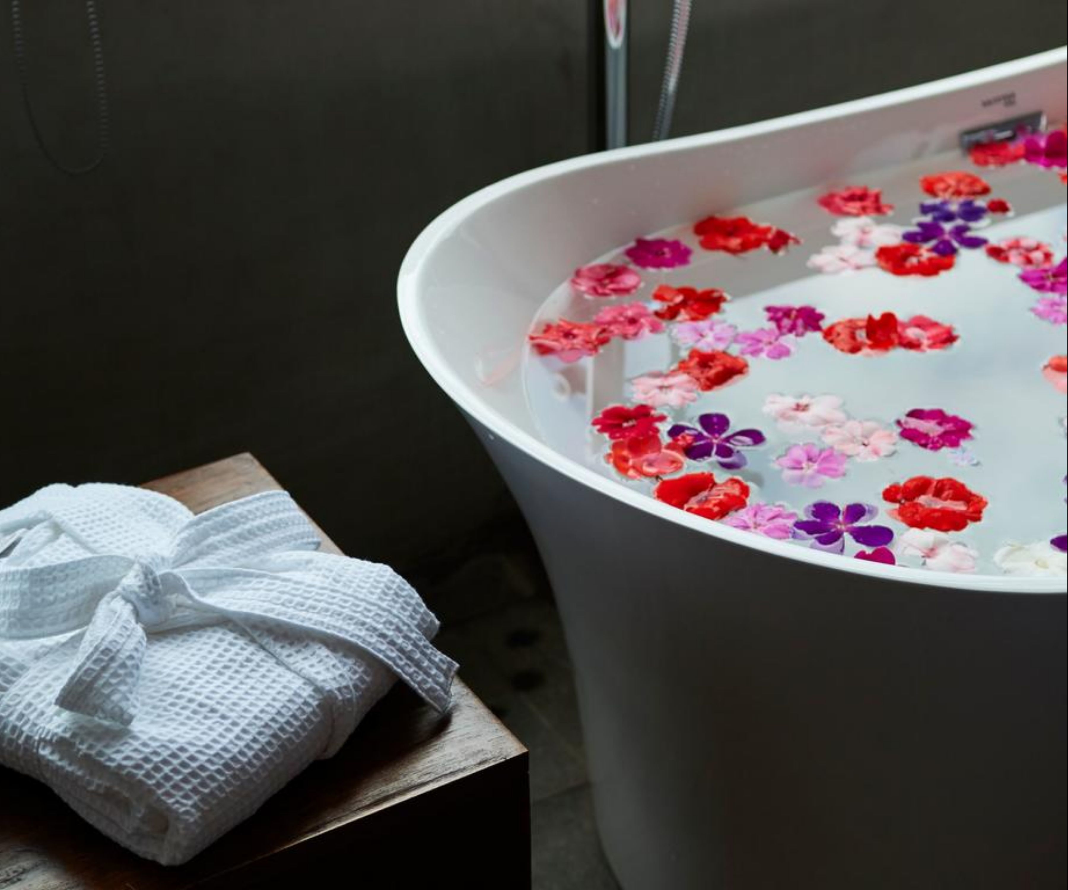 Une baignoire remplie d’eau avec des fleurs flottantes, une serviette blanche et une surface en bois.