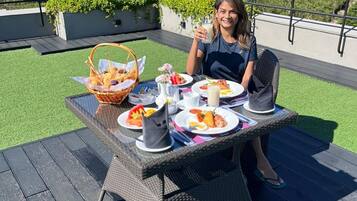 A dining table with plates of food, a basket, and a glass of drink on an outdoor deck with a view of green hills.