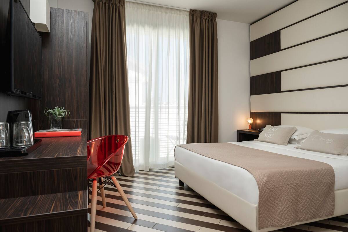 Een moderne hotelkamer met een bed, nachtkastje, lamp en een rode stoel.
