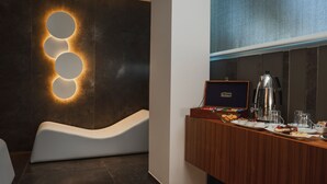 Un moderno vestíbulo de hotel con una zona de estar blanca y curva, una estación de café con una máquina de café de marca, y una pared con lámparas circulares.