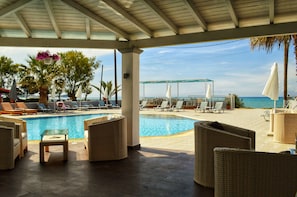 Uma área à beira da piscina com espreguiçadeiras, uma mesa pequena e um espaço coberto com vista para o mar.