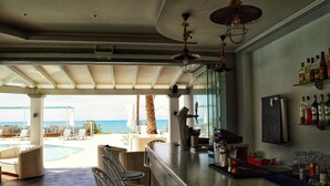 A bar area with a pool view, bar stools, and a bar counter with various bottles and glasses. - SunRose Beach Aparthotel (Corfu)