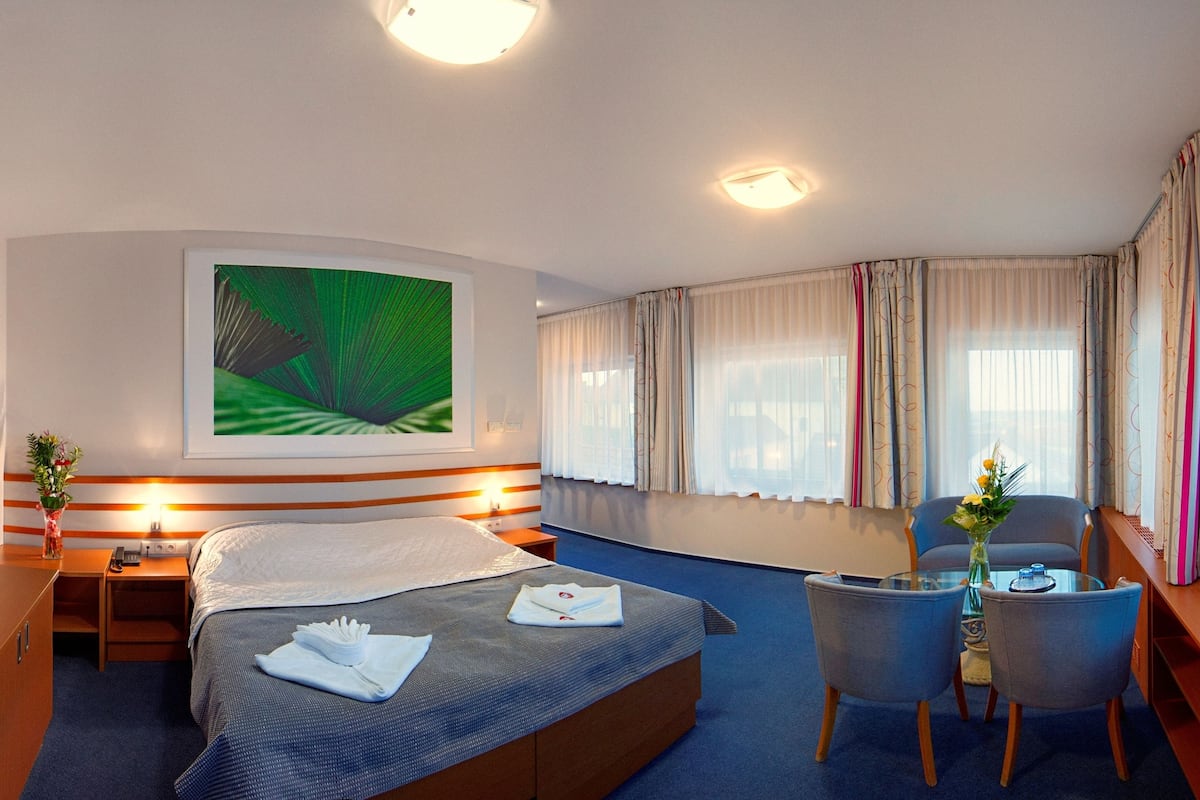 A hotel room with a bed, bedside tables, a desk, a chair, a small table with a vase, and curtains.