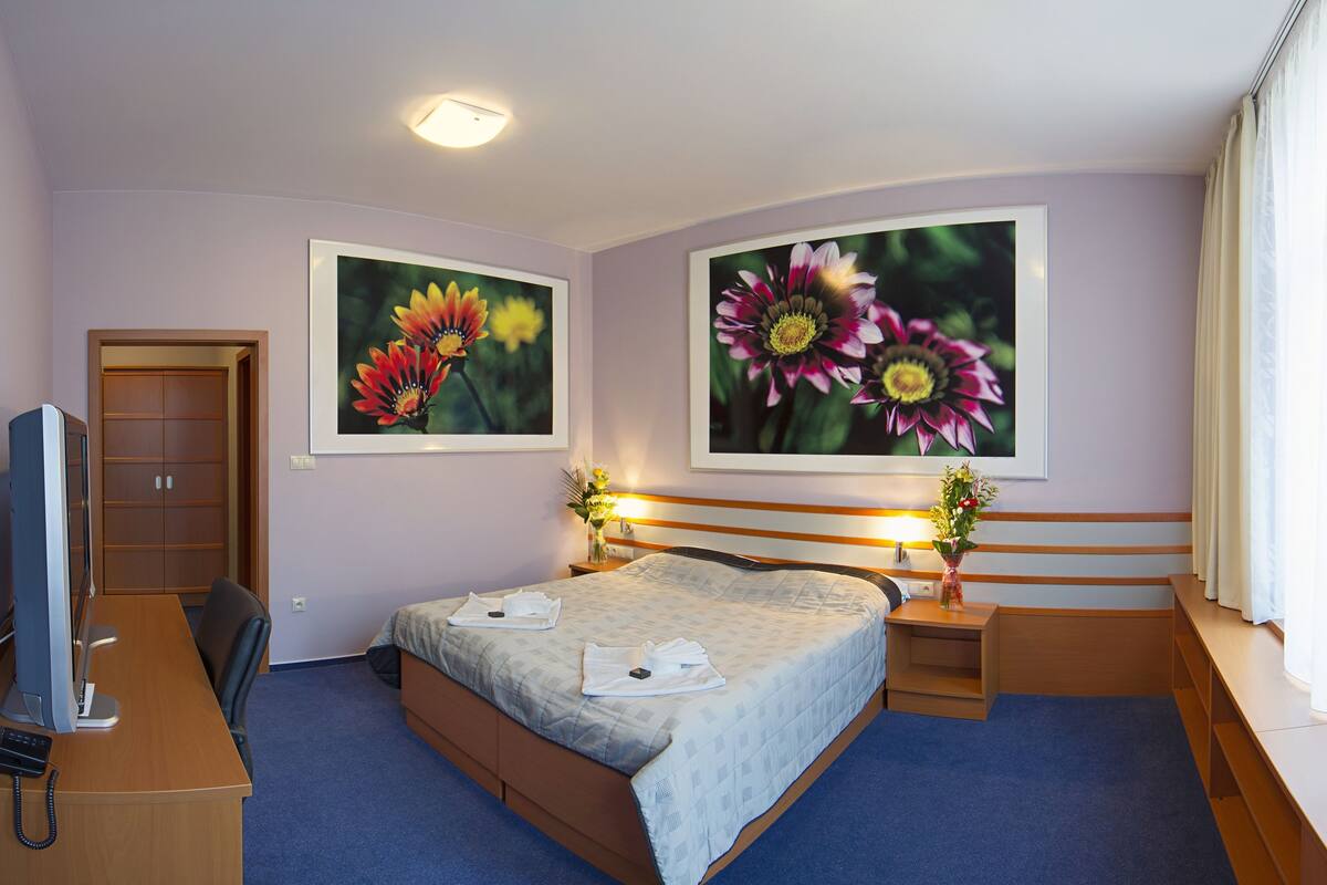 A hotel room with a bed, bedside tables, a desk with a computer, and two framed floral paintings on the wall.