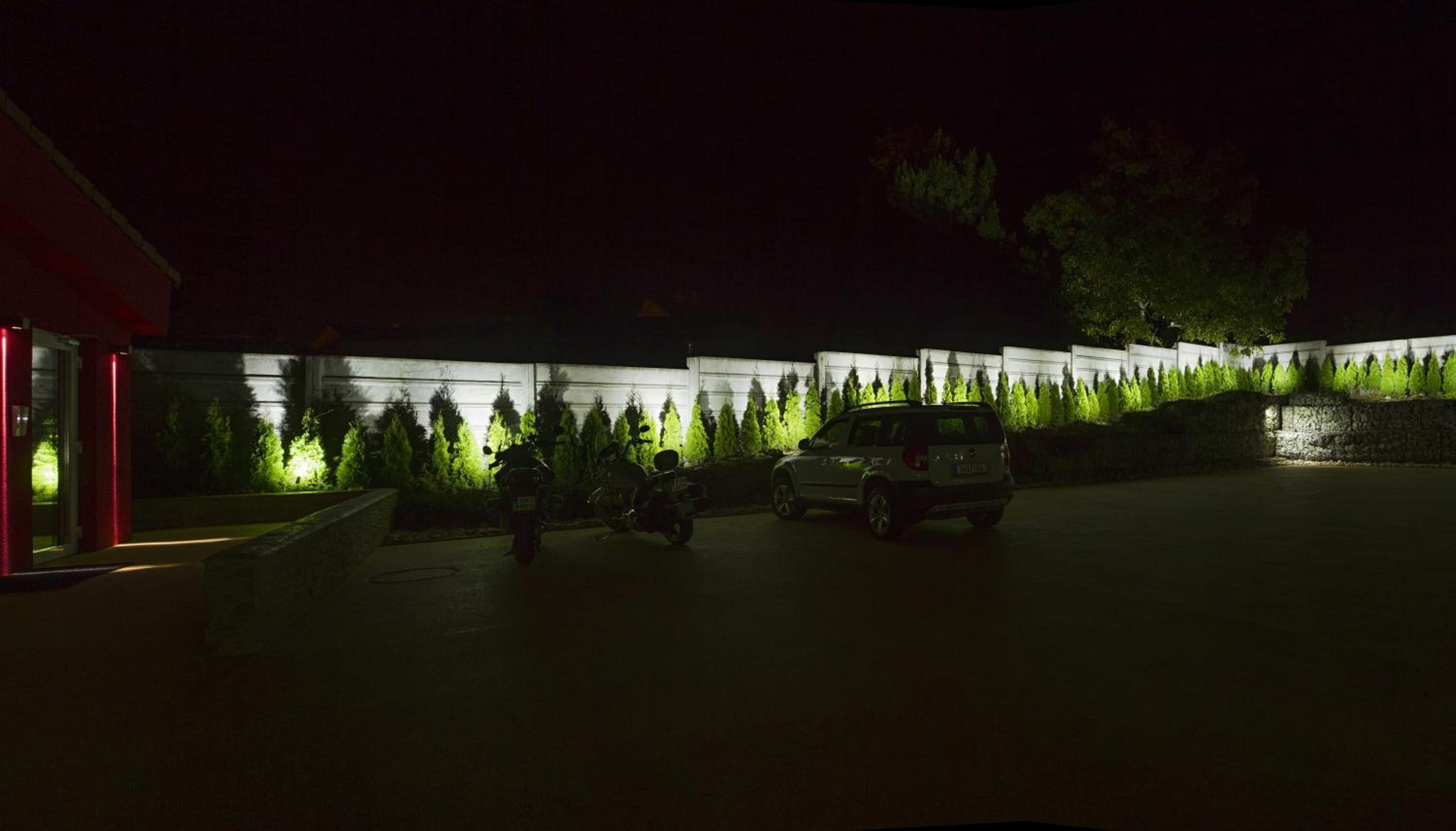 A parking area with two motorcycles and a car, surrounded by a low wall with green plants.