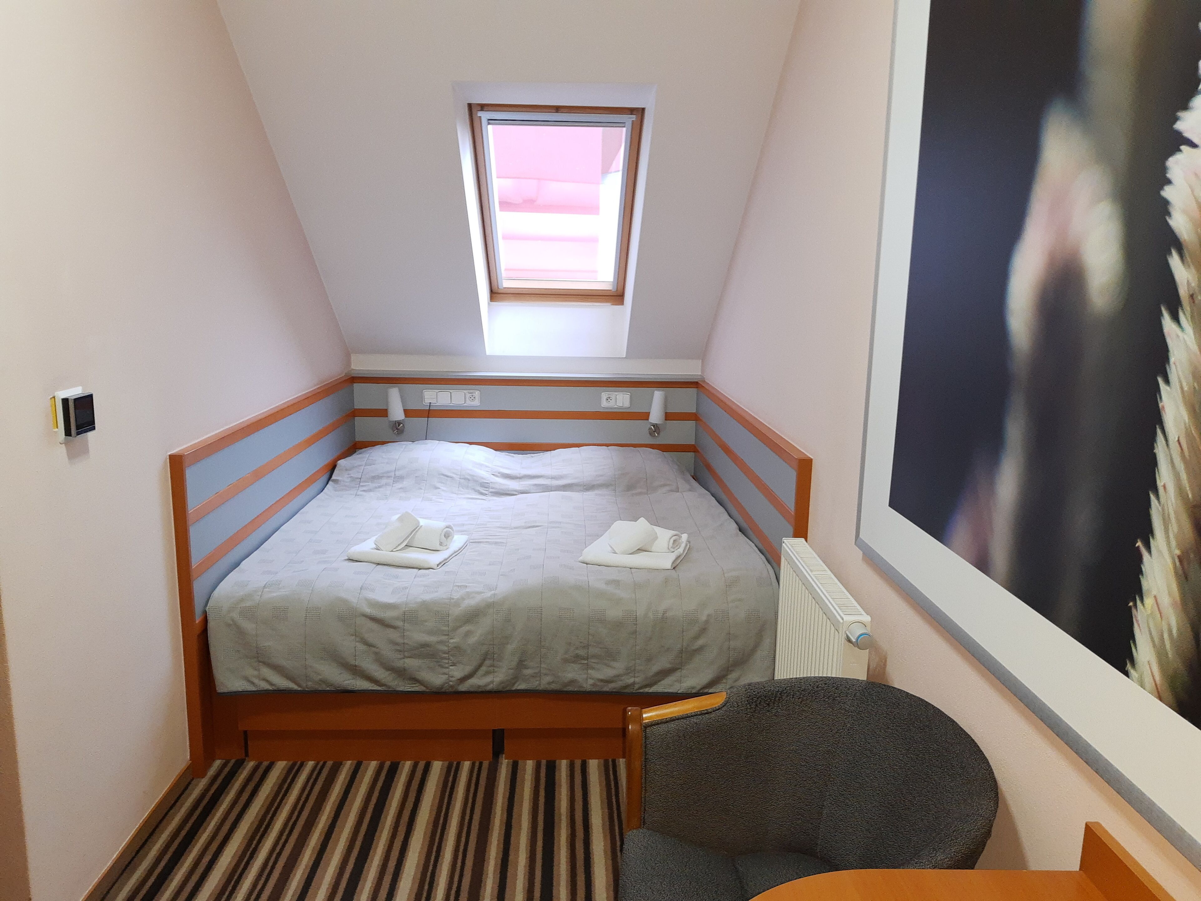 Quarto pequeno com cama de solteiro, teto inclinado, janela, radiador e uma cadeira.