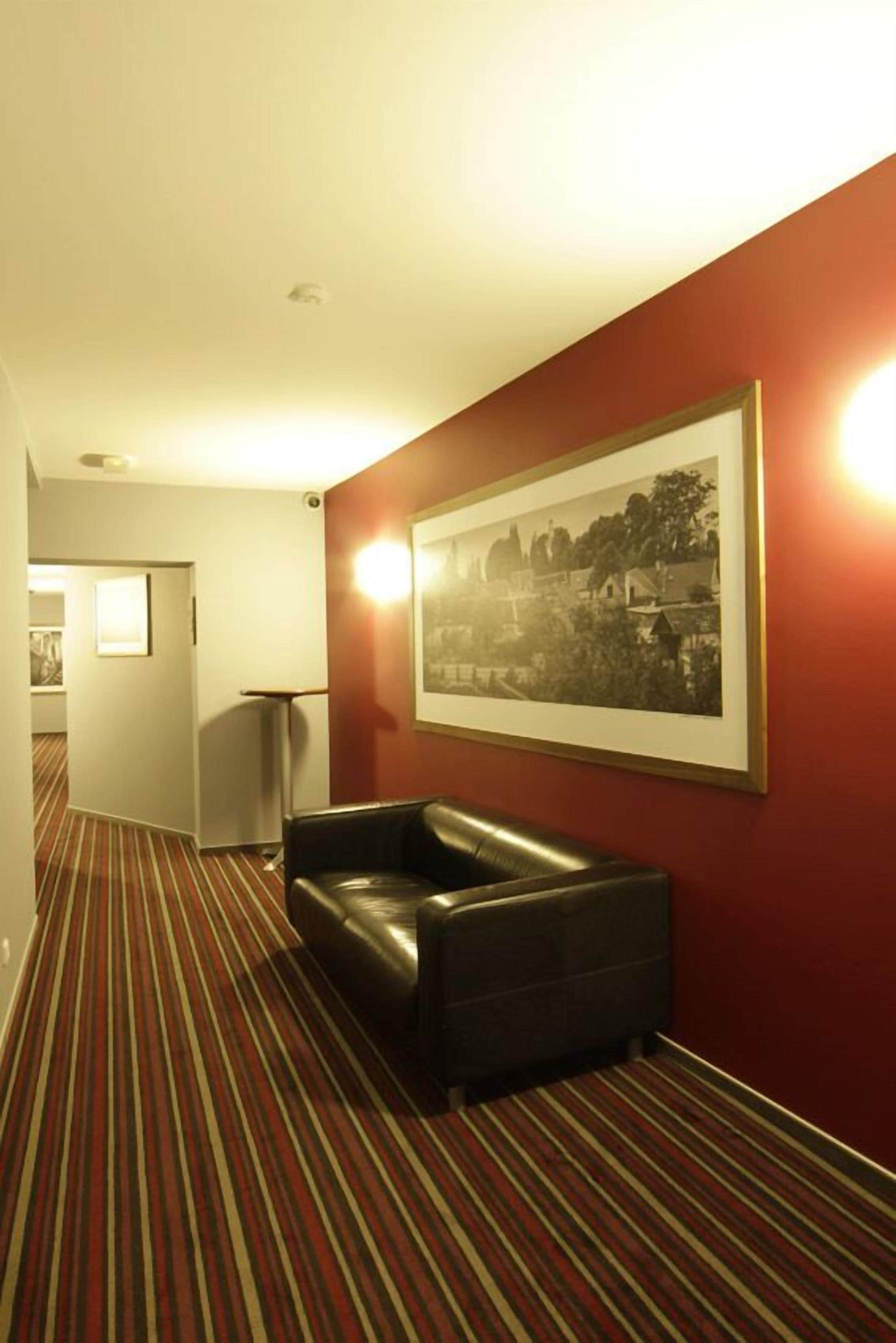 Um hall de hotel com carpete riscado, um sofá de couro preto e uma fotografia em preto e branco emoldurada na parede.