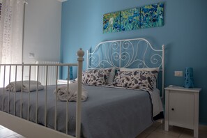 A bedroom with a bed, white metal headboard, patterned pillows, a small bedside table, and a blue wall with a floral painting. - My Room in Trani (Trani)