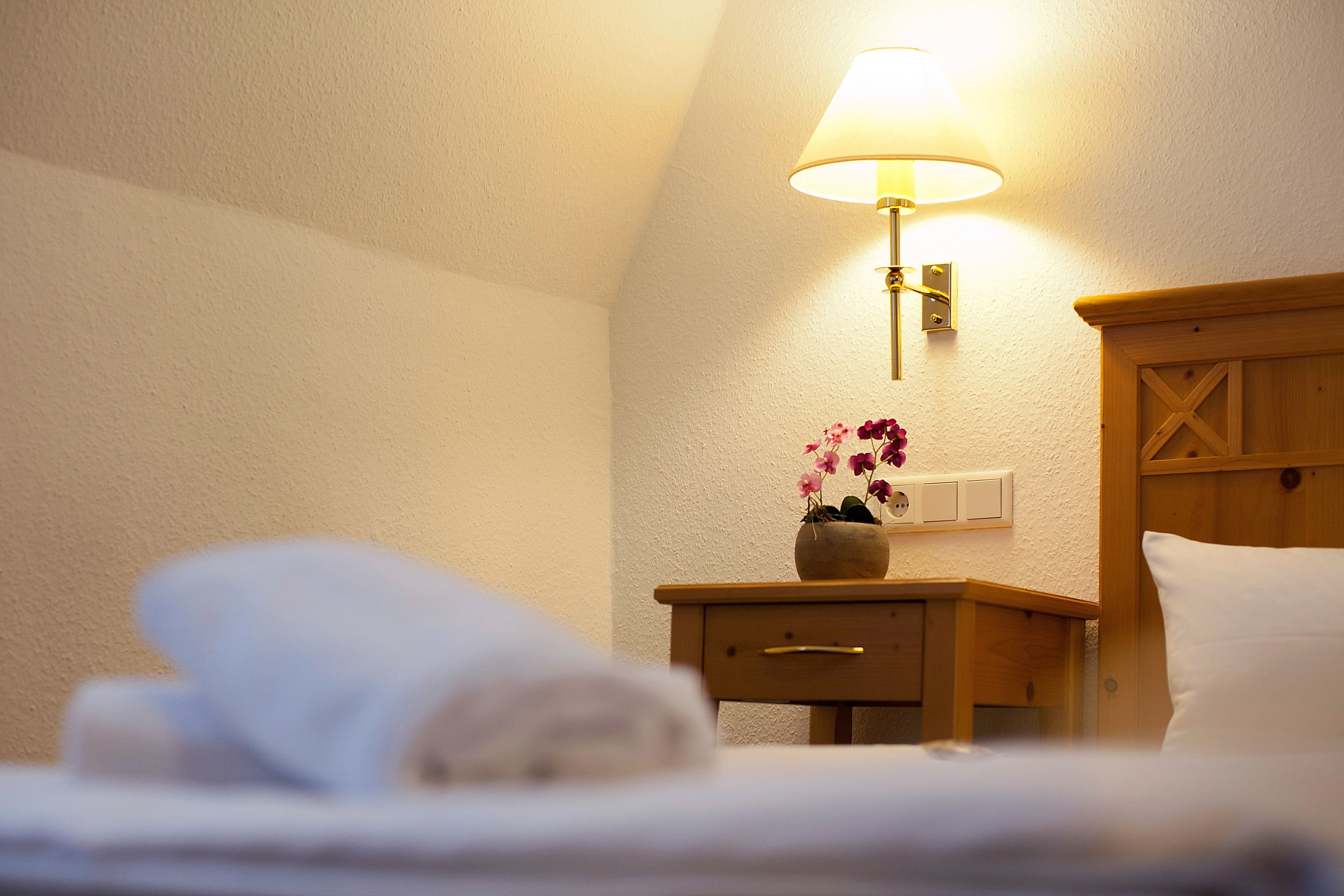Una cama bien hecha con una almohada blanca y una toalla doblada, una mesita de noche con una lámpara y una pequeña planta en maceta, y un cabecero de madera.