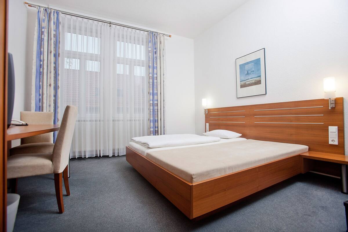 A hotel room with a wooden bed, a desk with a chair, and a window with curtains.