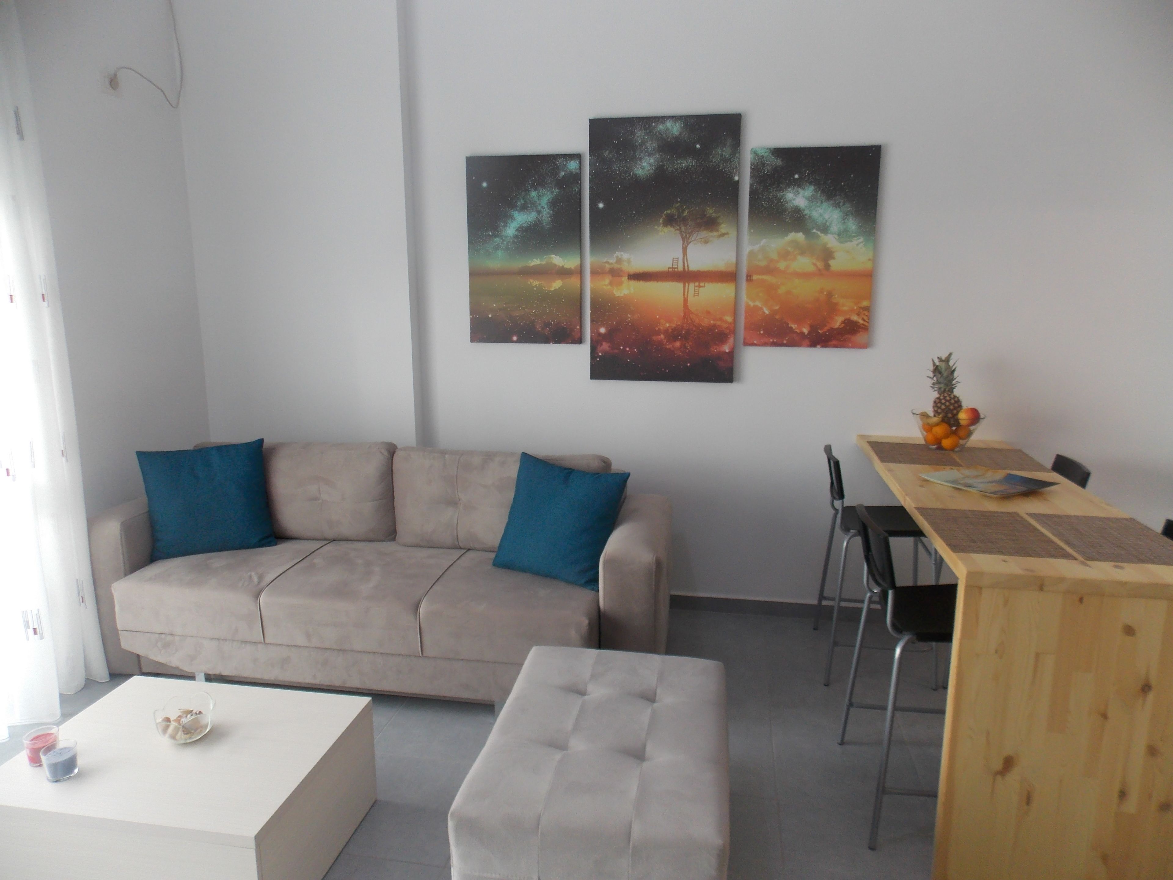 A modern living room with a beige sofa, two blue cushions, a white coffee table with candles, and a wooden dining table with a fruit bowl.