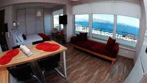 Habitación de hotel moderna con una mesa de comedor de madera, cojines rojos, un sofá y un amplio ventanal con vistas al mar.