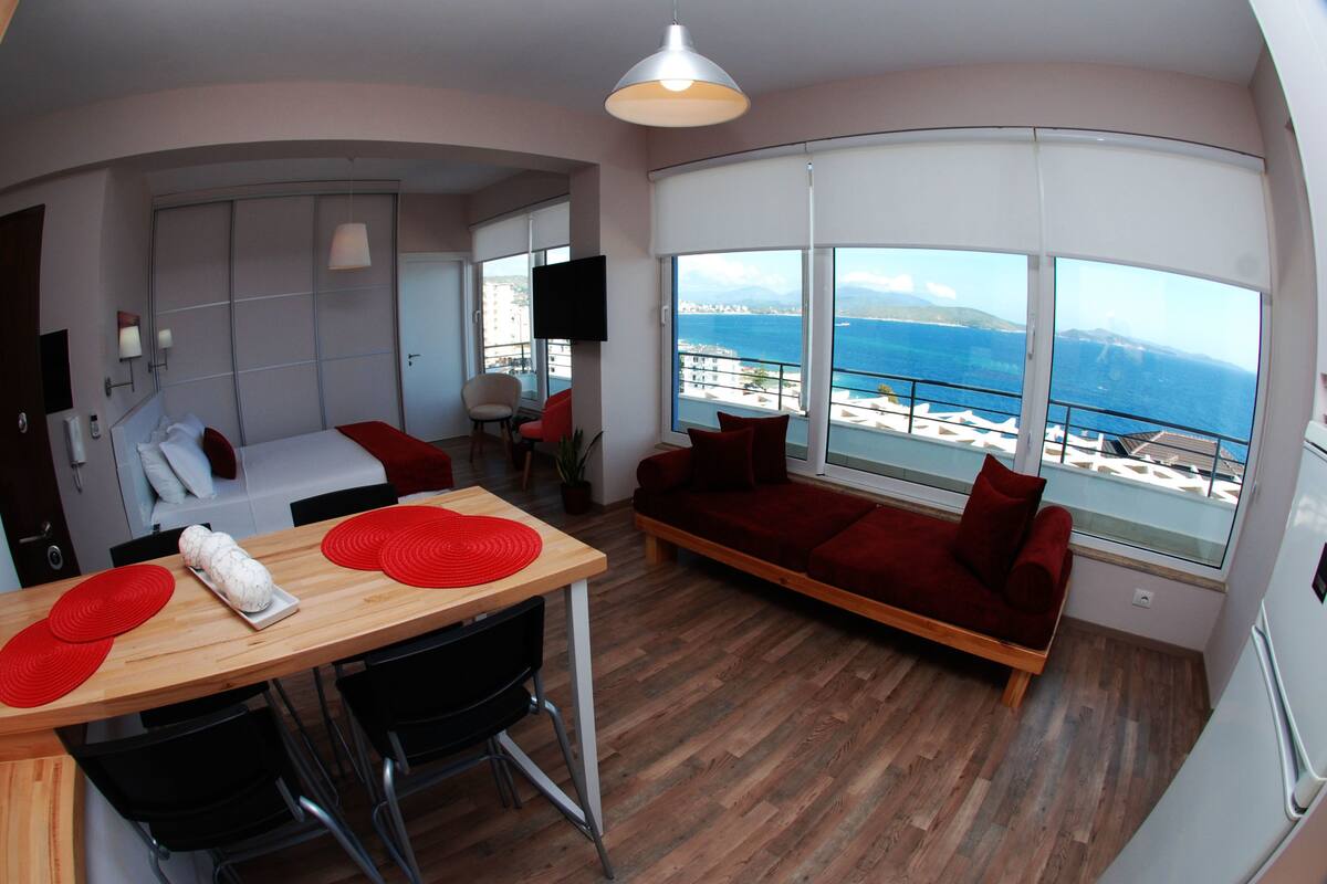 Habitación de hotel moderna con una mesa de comedor de madera, cojines rojos, un sofá y un amplio ventanal con vistas al mar.