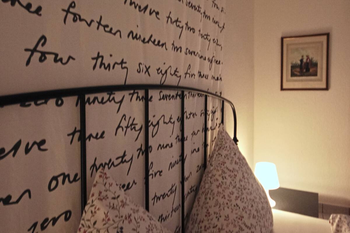 Une chambre avec un mur recouvert de chiffres écrits à la main, un lit orné d’oreillers à motifs floraux et un tableau encadré accroché au mur.