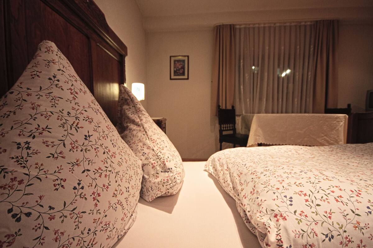 Une chambre d’hôtel avec deux lits, une literie à motifs floraux, une tête de lit en bois, une table de chevet avec une lampe, une chaise et une fenêtre avec des rideaux.