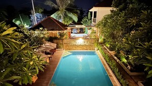 A swimming pool with a wooden deck, surrounded by tropical plants and a small building with a thatched roof. - Lavella Villa's (Kuta)