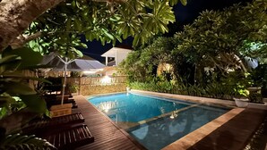 A swimming pool with a wooden deck and lounge chairs, surrounded by lush greenery and a stone wall. - Lavella Villa's (Kuta)