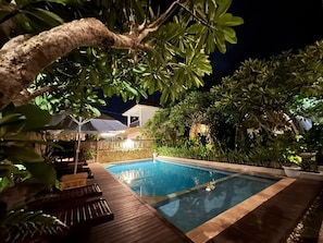 A swimming pool with a wooden deck and lounge chairs, surrounded by lush greenery and a stone wall. - Lavella Villa's (Kuta)