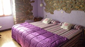 A bedroom with a stone wall, a bed with purple bedding, and a small bedside table. - Posada Elbete (Baztan)