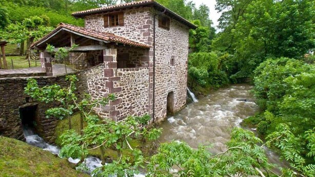 A stone building with a tiled roof, adjacent to a flowing river, surrounded by lush greenery. - Posada Elbete (Baztan)