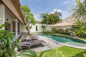 A poolside area with lounge chairs and a table, surrounded by greenery and a building with a thatched roof. - Villa Damai Taman (Seminyak)