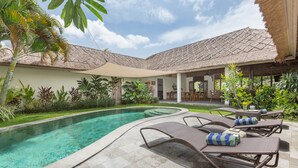 Un área junto a la piscina con sillas de descanso, una estructura con techo de paja y abundante vegetación.