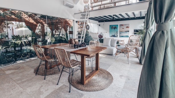 Un espacio habitable amplio con una mesa de comedor de madera, sillas de mimbre y un sofá. Se ve una escalera y una hamaca al fondo.