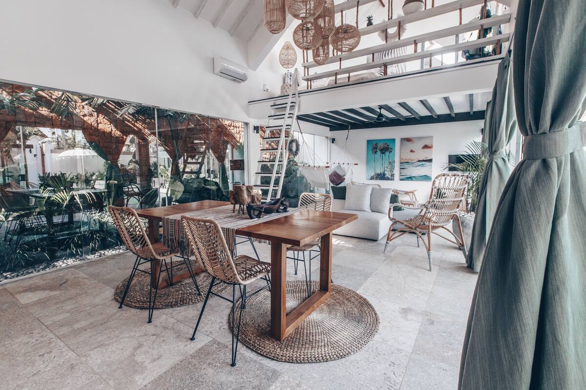 A spacious living area with a wooden dining table, wicker chairs, and a sofa. A ladder and a hammock are visible in the background.