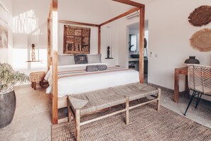 A four-poster bed with a woven bench, a potted plant, and a woven rug in a bright room with wooden beams and a large wall tapestry. - Beyond Bungalows (Canggu)