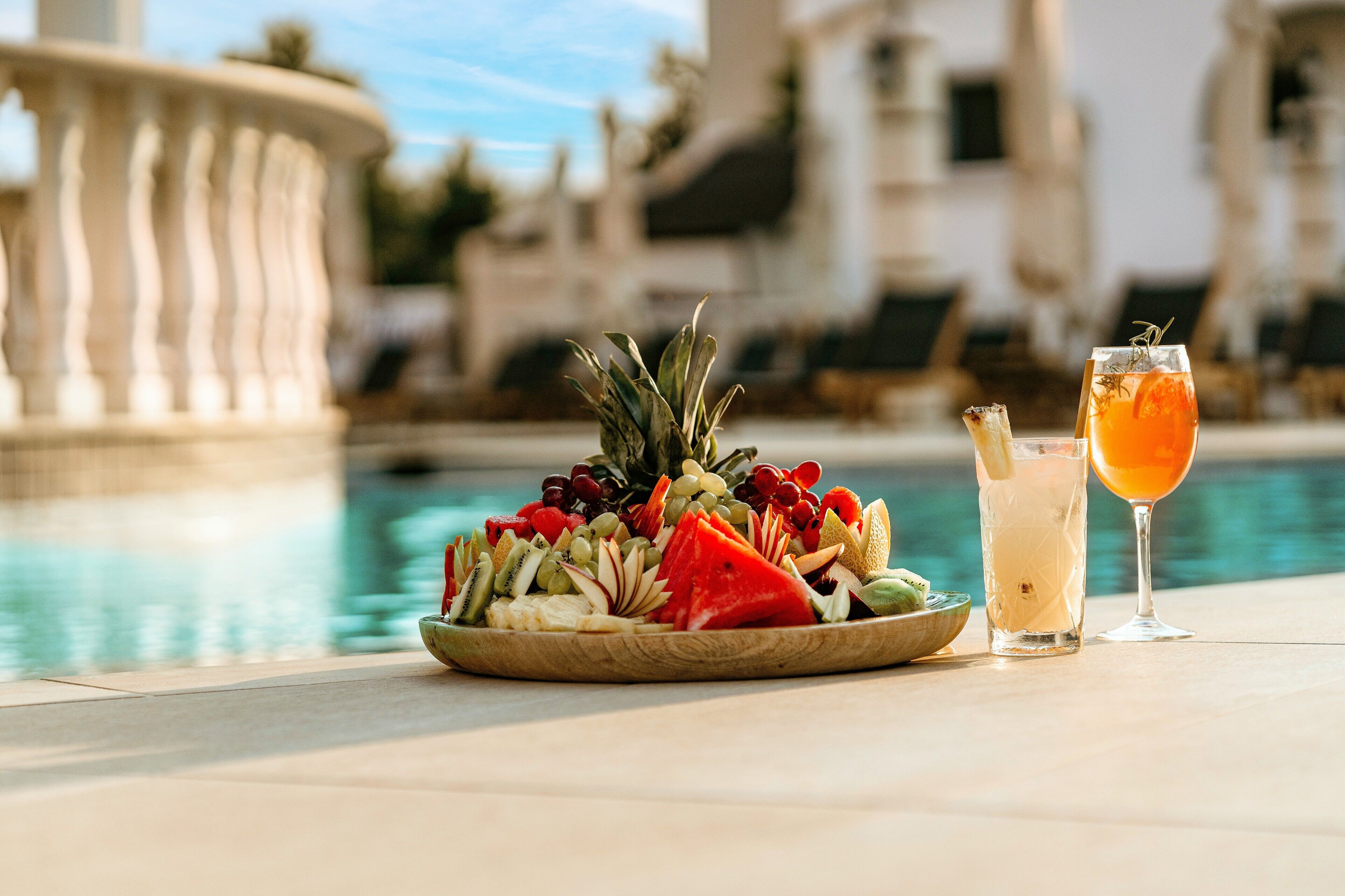 A fruit platter and drinks by a poolside with a fountain in the background.