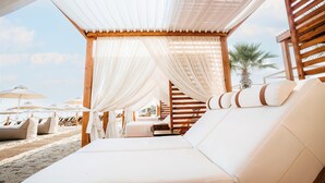 Um espaço de relaxamento à beira-mar com assentos estofados brancos, corrimãos de madeira e cortinas brancas.
