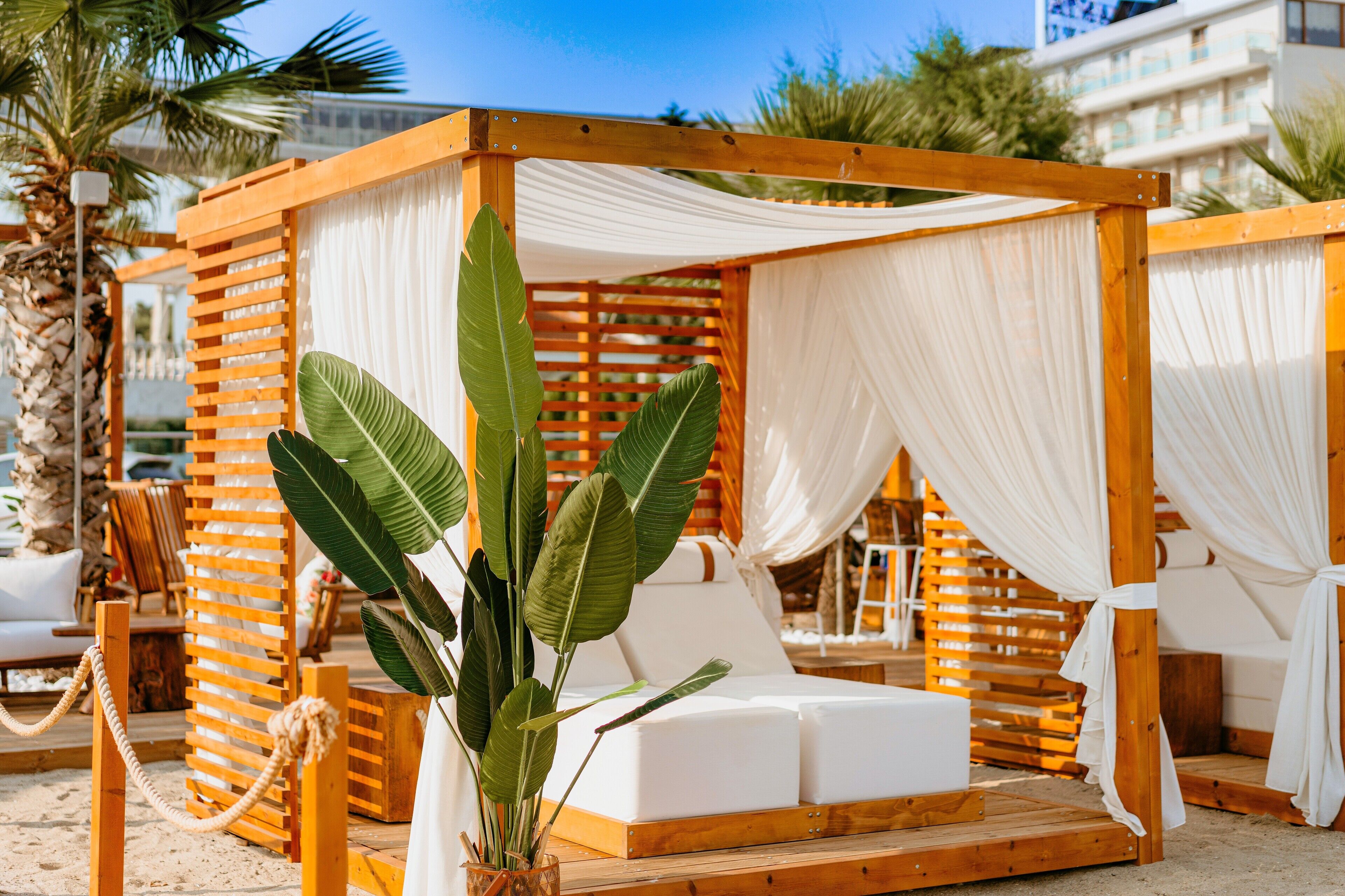 A wooden cabana with white cushions and a plant, set on a sandy beach with palm trees and a building in the background.