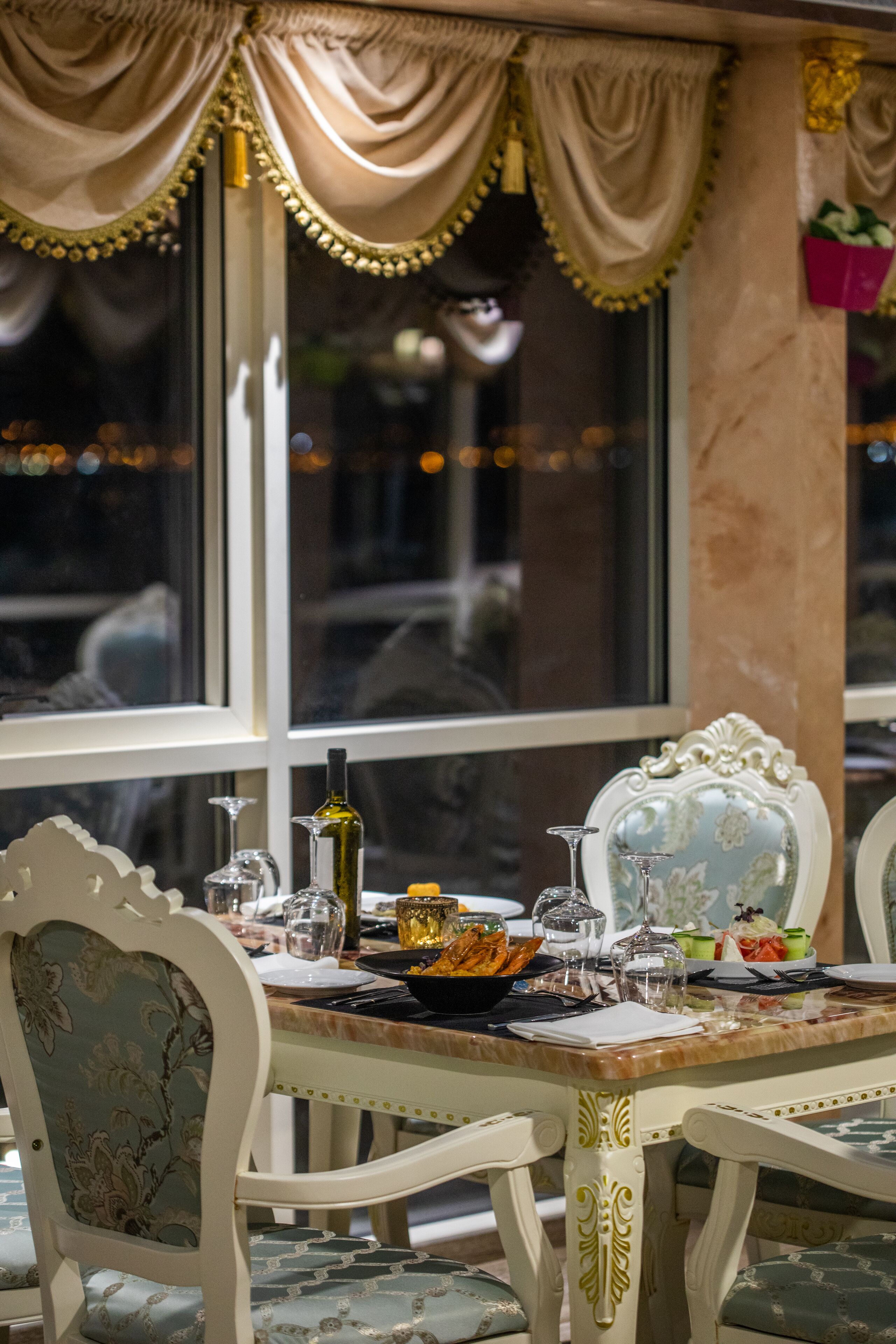 Una mesa de comedor dispuesta con copas y platos, rodeada de sillas ornamentadas con tapicería floral, frente a una ventana con cortinas.
