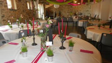 Une salle de banquet avec des tables rondes dressées pour un événement formel, agrémentée de nappes blanches, de serviettes rouges et de petits plants en pot.