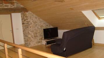 A small attic room with a stone wall, a black sofa, a television, and a wooden ceiling.