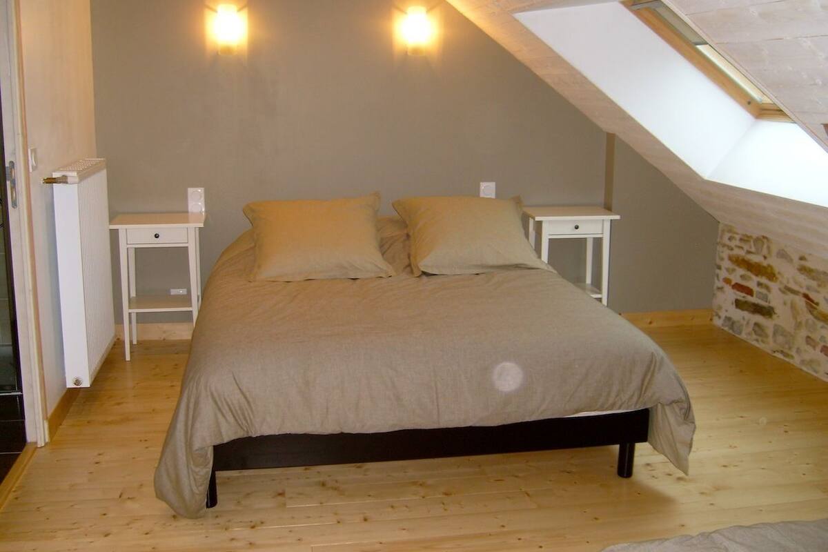 Quarto com teto inclinado, uma cama com duas almofadas, duas mesas de cabeceira e um candeeiro fixado na parede.