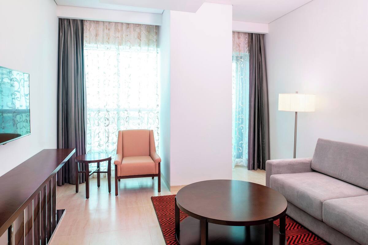 A modern hotel room with a sofa, armchair, round coffee table, and a TV on a stand.