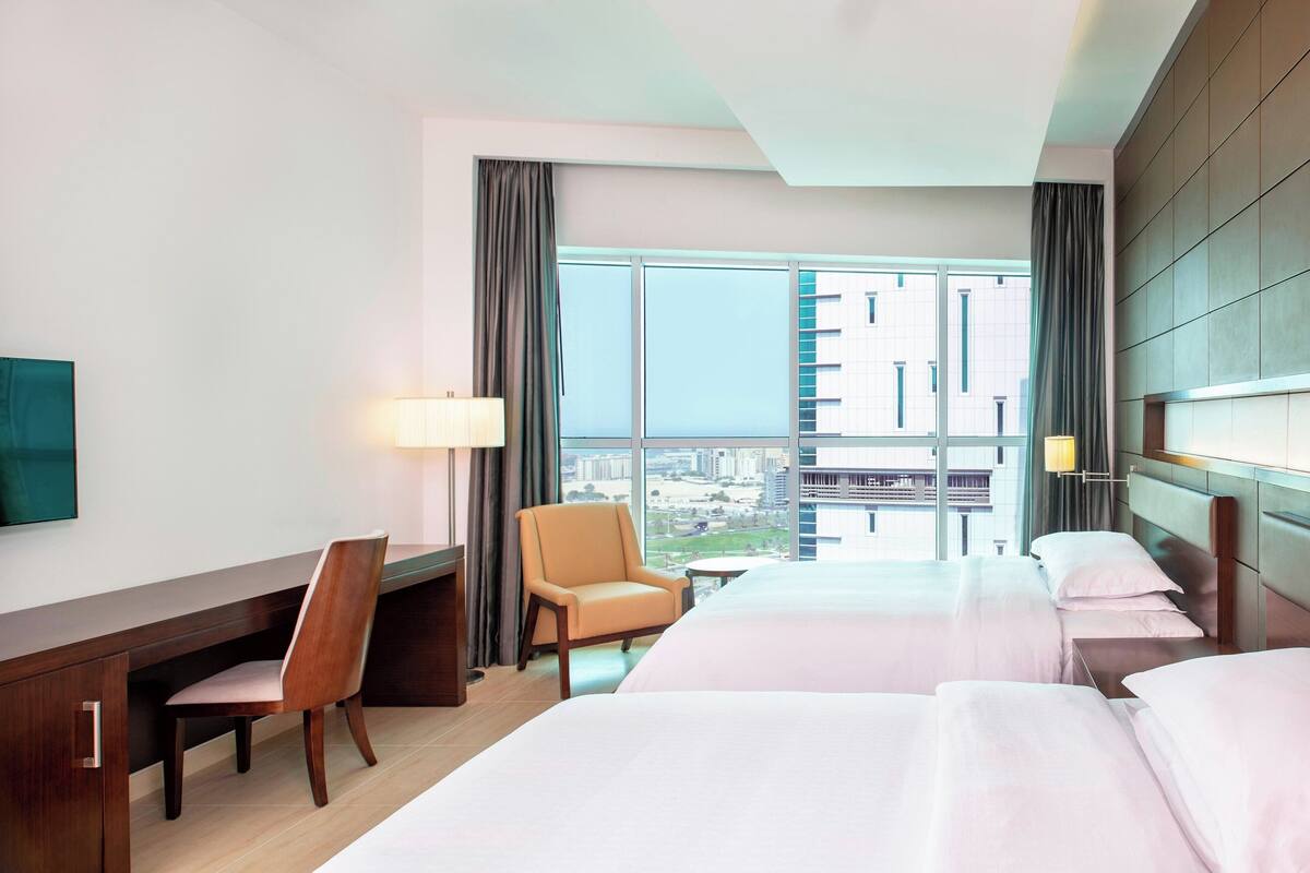 A hotel room with a desk, a chair, a bed, and a view of the cityscape through large windows.
