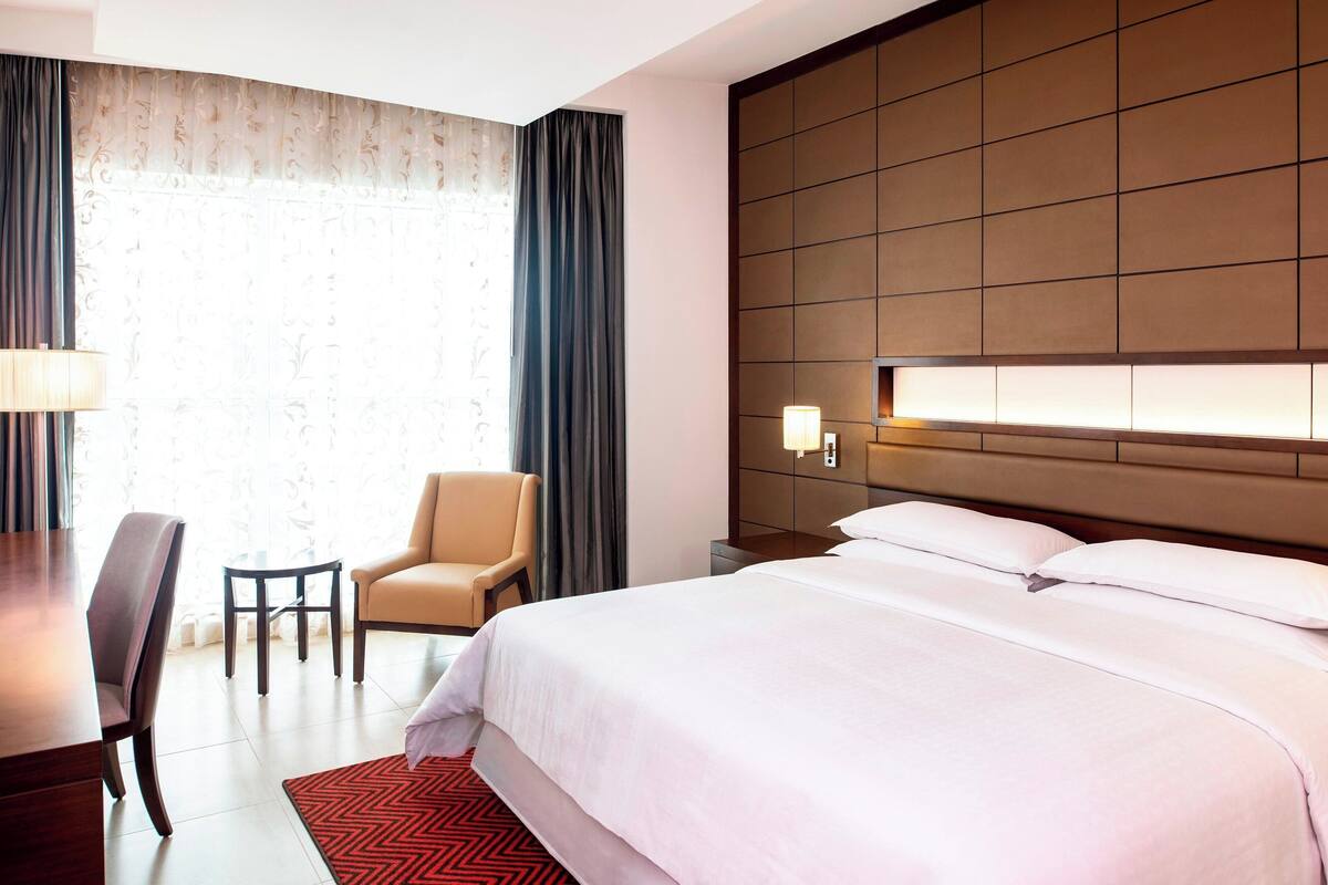 A hotel room with a large bed, a desk, a chair, and a nightstand with a lamp.