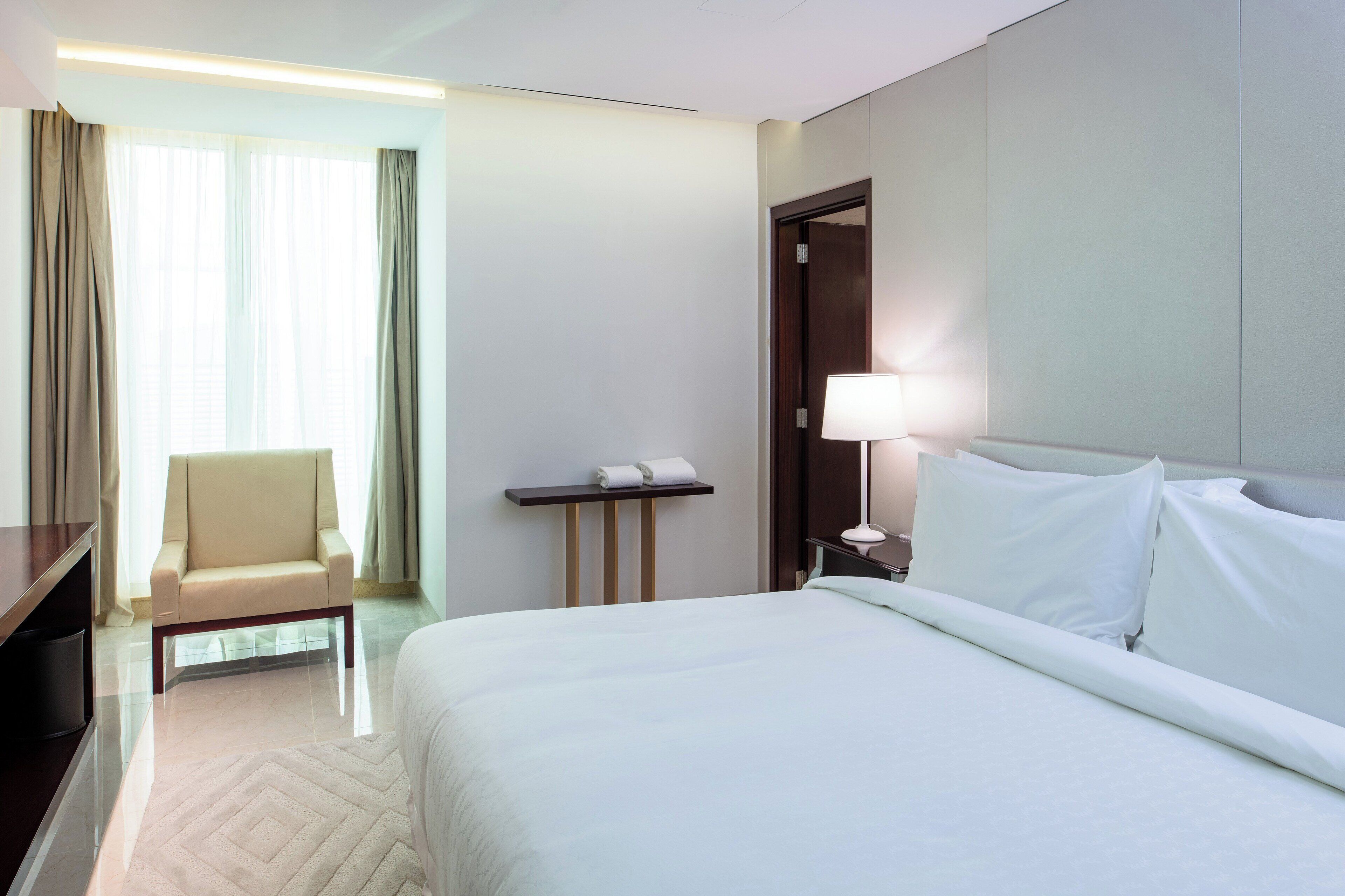 A hotel room with a bed, a chair, a desk, and a lamp.