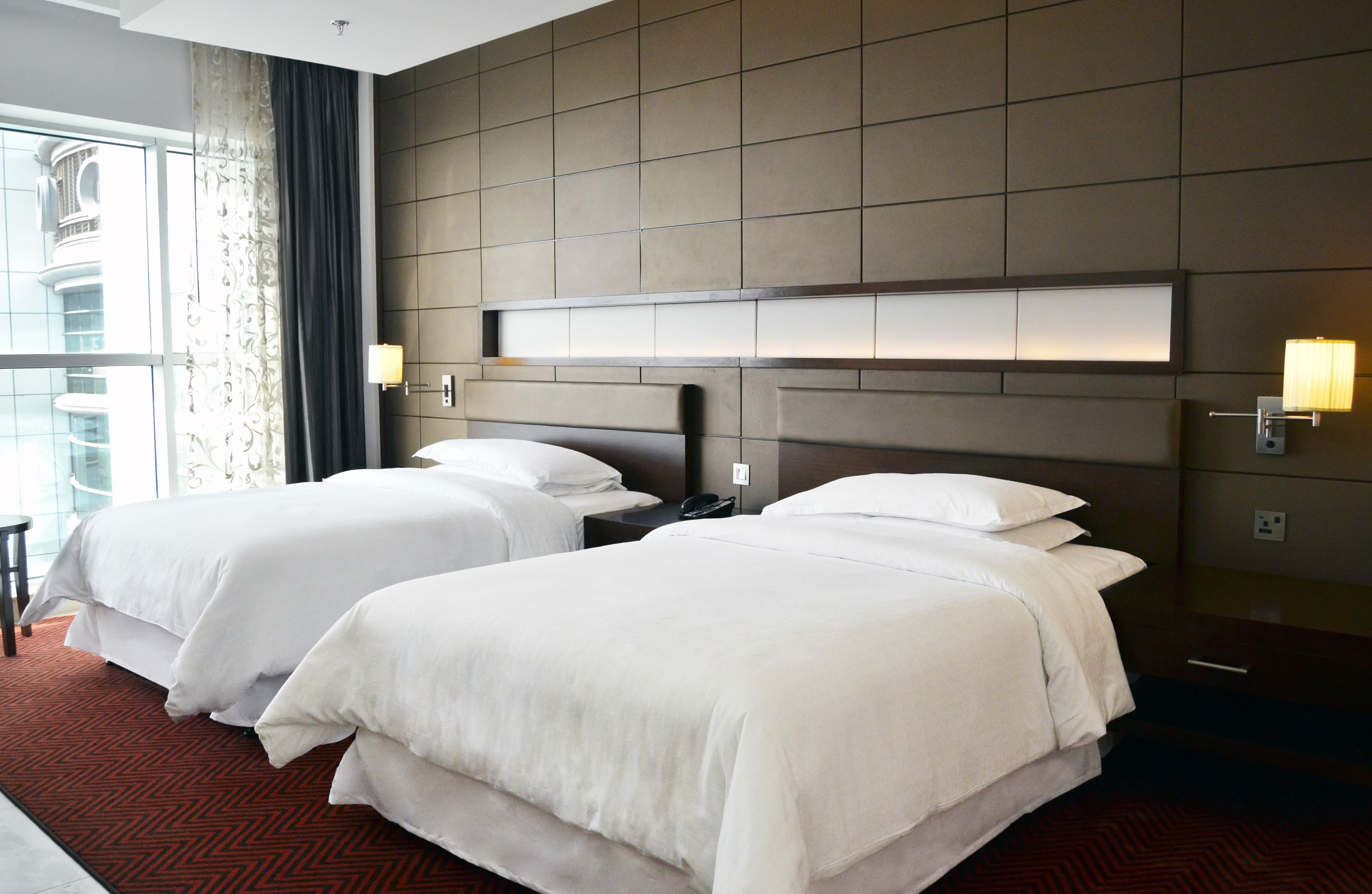 A hotel room with two beds, a large window with curtains, a modern headboard, and wall-mounted lamps.
